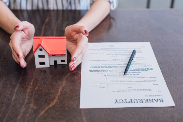 Partial view of female hands near house model, paper with bankruptcy lettering and pen on table